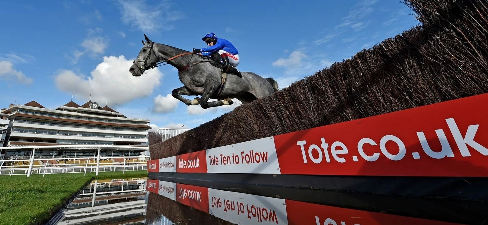A wide-angle photograph capturing a horse and jockey mid-air as they leap over a water jump.